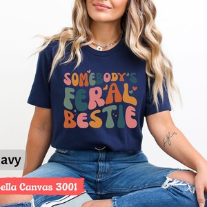 May include: A navy blue t-shirt with the text "Somebody's Feral Bestie" in colorful, retro-style lettering. The shirt is made by Bella Canvas and is a size 3001.