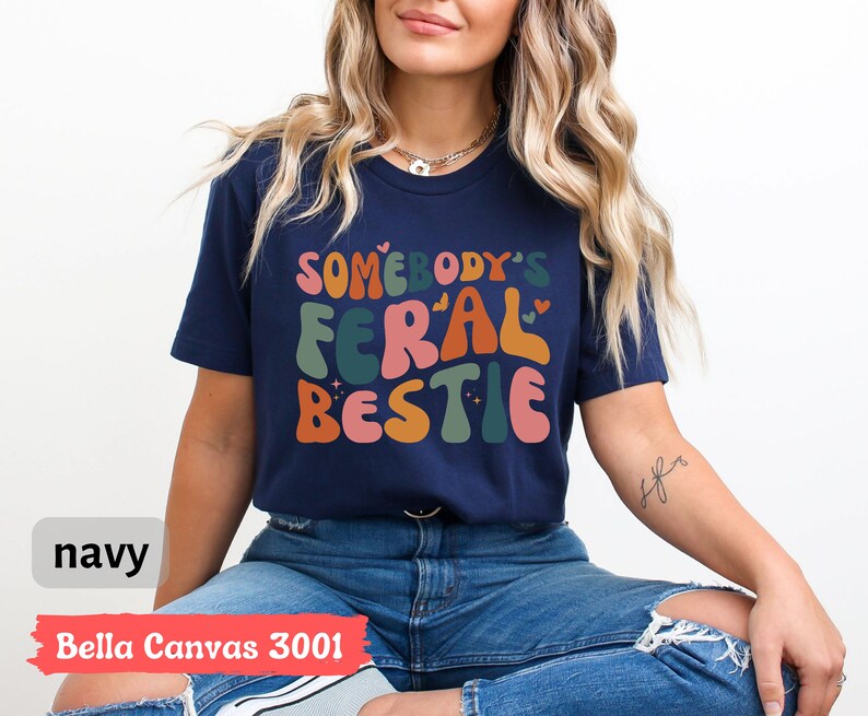 May include: A navy blue t-shirt with the text "Somebody's Feral Bestie" in colorful, retro-style lettering. The shirt is made by Bella Canvas and is a size 3001.