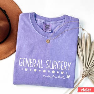 Może przedstawiać: Fioletowy T-shirt z napisem "GENERAL SURGERY nurse" w białym kolorze z małym sercem i akcentami w postaci stokrotek. Koszulka jest złożona i leży płasko na białym tle.