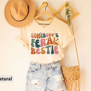 May include: A beige t-shirt with the text "Somebody's Feral Bestie" in colorful, retro-style lettering. The shirt is hanging on a wooden hanger with a straw bag and a hat.
