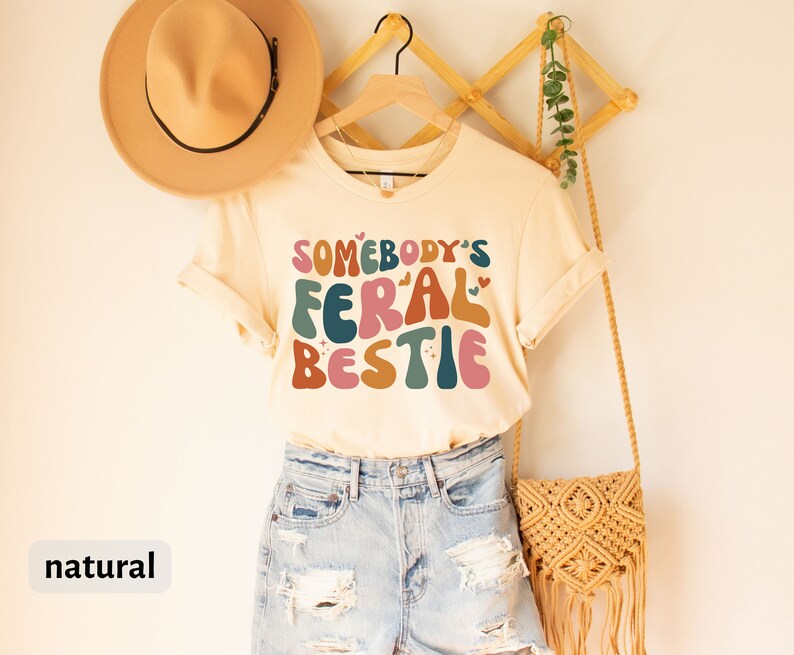 May include: A beige t-shirt with the text "Somebody's Feral Bestie" in colorful, retro-style lettering. The shirt is hanging on a wooden hanger with a straw bag and a hat.