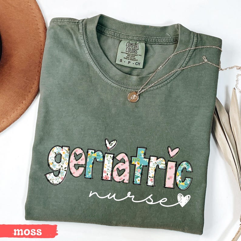 Pode incluir: Camiseta Comfort Colors verde musgo com as palavras "geriatric nurse" em um design floral. Um delicado colar de ouro repousa sobre a camiseta. A camiseta &eacute; dobrada e exibida com um chap&eacute;u marrom e folhas secas.