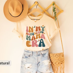 May include: A beige t-shirt with the text "In My Homeschool Mama Era" in colorful, retro-style lettering. The shirt is hanging on a wooden rack with a straw bag and a hat.
