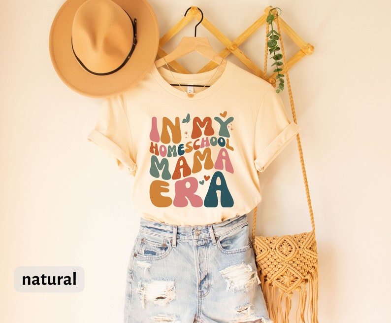 May include: A beige t-shirt with the text "In My Homeschool Mama Era" in colorful, retro-style lettering. The shirt is hanging on a wooden rack with a straw bag and a hat.