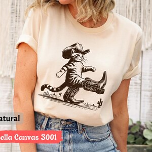 May include: A natural-colored t-shirt featuring a cartoon cat wearing a cowboy hat and boots. The cat is walking with a confident stride. The text "Bella Canvas 3001" is printed on the bottom of the shirt.