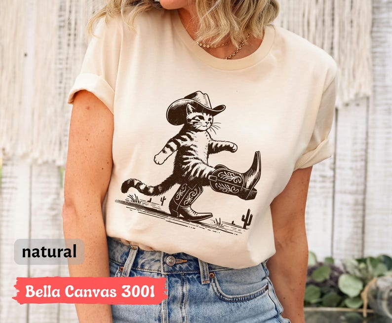 May include: A natural-colored t-shirt featuring a cartoon cat wearing a cowboy hat and boots. The cat is walking with a confident stride. The text "Bella Canvas 3001" is printed on the bottom of the shirt.