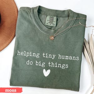 May include: A moss green t-shirt with the text "helping tiny humans do big things" in white. A small white heart graphic is below the text. A gold necklace with a pendant is on the shirt. The shirt is labeled "Comfort Colors".