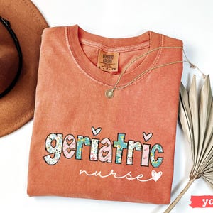Pode incluir: Uma camiseta Comfort Colors cor inhame com as palavras "Geriatric Nurse" em um design floral e de cora&ccedil;&atilde;o. Um colar de ouro e um chap&eacute;u marrom est&atilde;o ao lado da camisa.