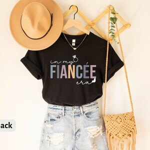 May include: Black t-shirt with the text "in my fianc&eacute;e era" in colorful letters with a ring and arrow graphic. The shirt is hanging on a wooden rack with a straw bag and a hat.