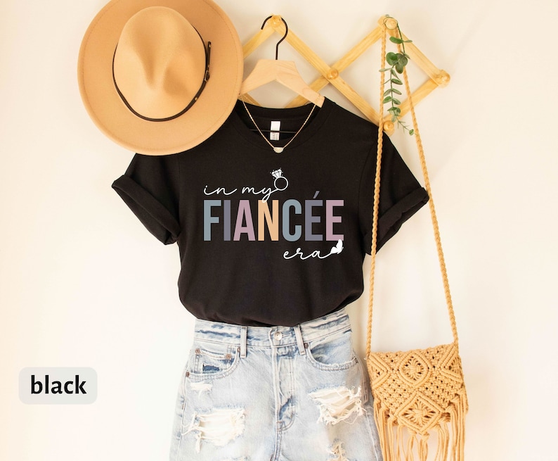 May include: Black t-shirt with the text "in my fianc&eacute;e era" in colorful letters with a ring and arrow graphic. The shirt is hanging on a wooden rack with a straw bag and a hat.