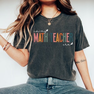 May include: A dark grey t-shirt with the text "in my MATH TEACHER era" in rainbow colours. The text is in a playful font with a heart and butterfly graphic.