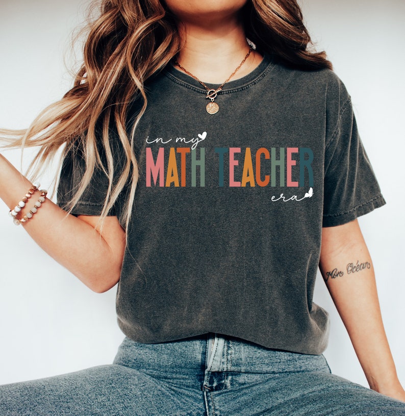 May include: A dark grey t-shirt with the text "in my MATH TEACHER era" in rainbow colours. The text is in a playful font with a heart and butterfly graphic.