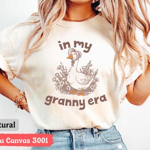 May include: A natural-colored t-shirt with the text "in my granny era" above a cartoon goose wearing a bonnet. The goose is surrounded by flowers. The shirt is Bella Canvas 3001.