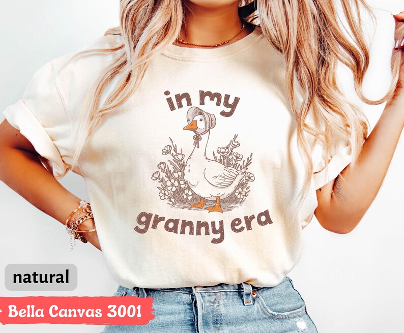 May include: A natural-colored t-shirt with the text "in my granny era" above a cartoon goose wearing a bonnet. The goose is surrounded by flowers. The shirt is Bella Canvas 3001.