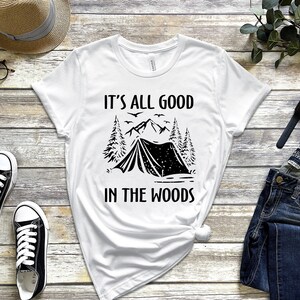 May include: White t-shirt with black text and graphic. The text reads "It's all good in the woods". The graphic is a tent in a forest with mountains in the background.