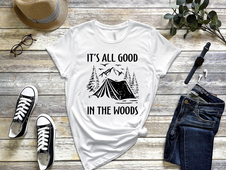 May include: White t-shirt with black text and graphic. The text reads "It's all good in the woods". The graphic is a tent in a forest with mountains in the background.