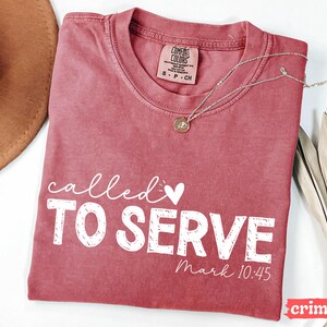 Pu&ograve; includere: T-shirt color cremisi con le parole "called TO SERVE" in scrittura bianca e lettere maiuscole. Il testo "Mark 10:45" &egrave; in basso. Una collana d'oro con un ciondolo &egrave; appoggiata sulla maglietta.