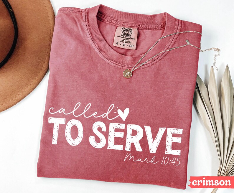 Pu&ograve; includere: T-shirt color cremisi con le parole "called TO SERVE" in scrittura bianca e lettere maiuscole. Il testo "Mark 10:45" &egrave; in basso. Una collana d'oro con un ciondolo &egrave; appoggiata sulla maglietta.