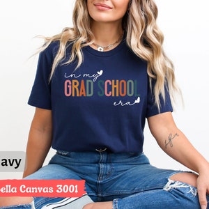 May include: A navy blue t-shirt with the text "in my grad school era" in colorful letters and white hearts. The shirt is made by Bella Canvas and is a 3001 style.