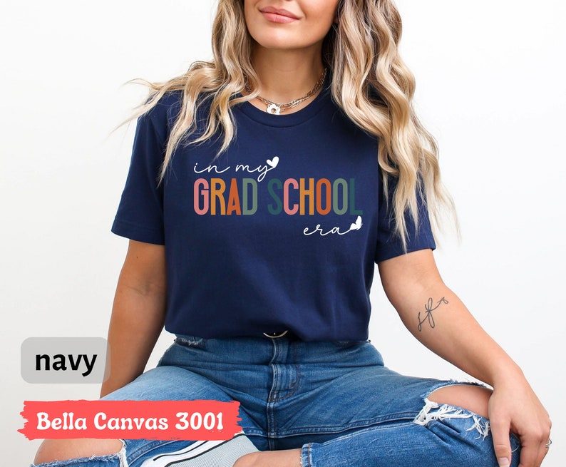 May include: A navy blue t-shirt with the text "in my grad school era" in colorful letters and white hearts. The shirt is made by Bella Canvas and is a 3001 style.