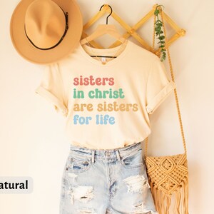 May include: A beige t-shirt with the text "sisters in christ are sisters for life" in colorful letters. The shirt is hanging on a wooden hanger with a straw hat and a woven bag.