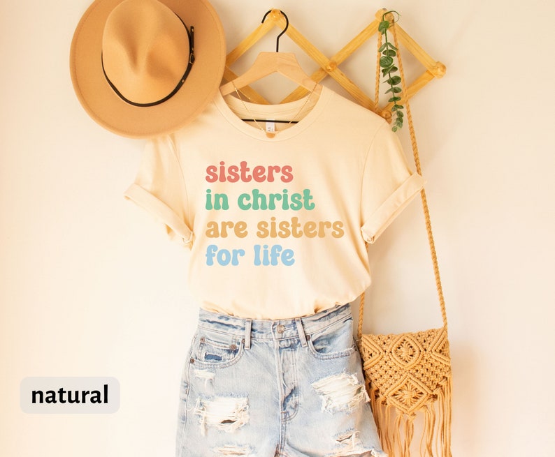 May include: A beige t-shirt with the text "sisters in christ are sisters for life" in colorful letters. The shirt is hanging on a wooden hanger with a straw hat and a woven bag.