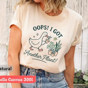May include: A cream-colored t-shirt with the text "OOPS! I GOT Another Plant!" and a graphic of a duck carrying a potted plant. The shirt is made of Bella Canvas 3001 material.