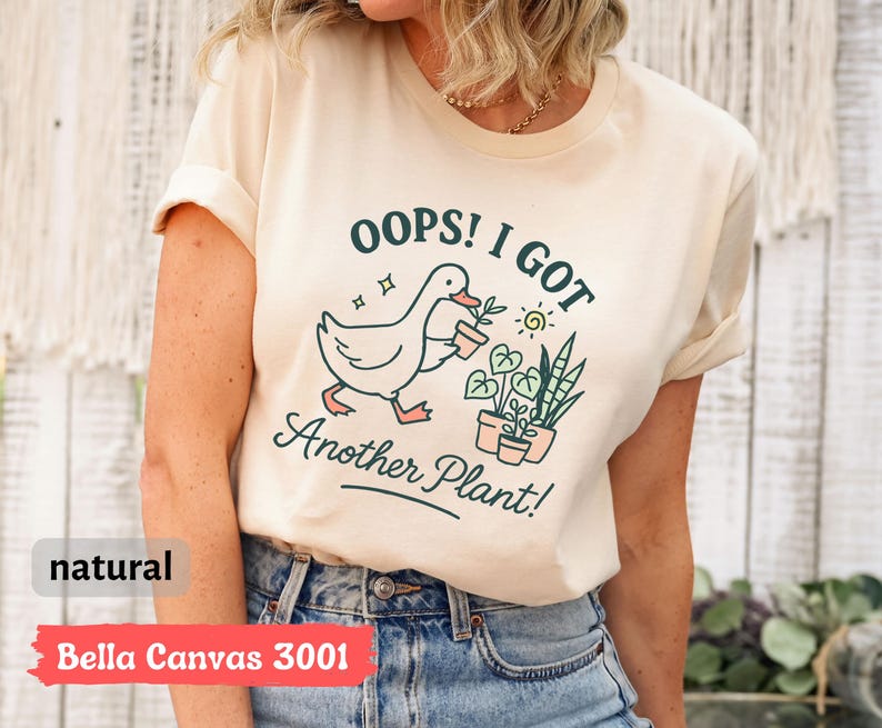 May include: A cream-colored t-shirt with the text "OOPS! I GOT Another Plant!" and a graphic of a duck carrying a potted plant. The shirt is made of Bella Canvas 3001 material.