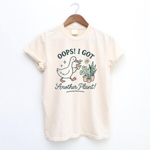 May include: Cream-colored t-shirt with a graphic of a duck carrying a potted plant, with the text "Oops! I Got Another Plant!" in green and black. The design includes potted plants and a sun. The shirt is hanging on a wooden hanger.