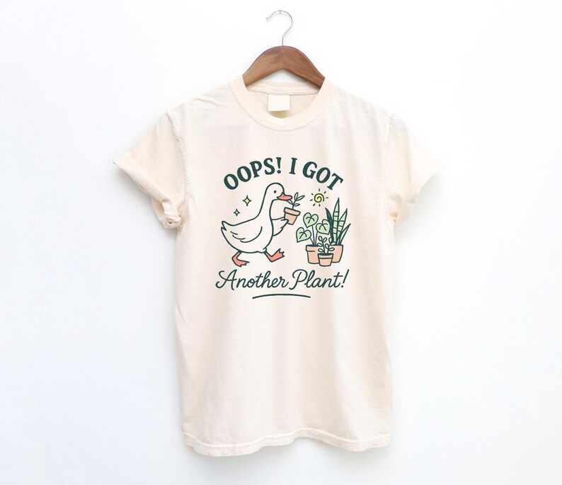 May include: Cream-colored t-shirt with a graphic of a duck carrying a potted plant, with the text "Oops! I Got Another Plant!" in green and black. The design includes potted plants and a sun. The shirt is hanging on a wooden hanger.