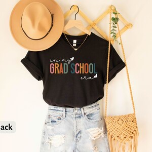 May include: Black t-shirt with the text "in my grad school era" in rainbow colors with a heart and butterfly graphic.
