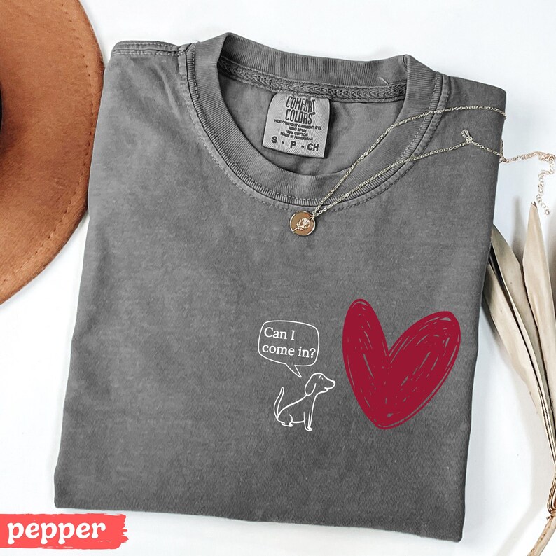 May include: A gray Comfort Colors t-shirt with a red heart and a cartoon dog with the text "Can I come in?". A gold necklace with a pendant is on the shirt. The word "pepper" is on a red banner.