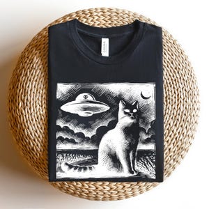 May include: Black t-shirt with a white graphic print of a cat looking up at a flying saucer. The graphic is in a vintage woodcut style.