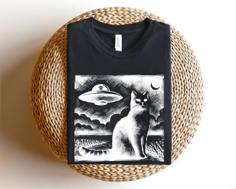 May include: Black t-shirt with a white graphic print of a cat looking up at a flying saucer. The graphic is in a vintage woodcut style.