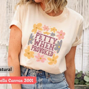 May include: A cream-colored t-shirt with the text "PETTY WHEN PROVOKED" in a square floral design. The design includes yellow, pink, and blue flowers. The shirt is made of Bella Canvas 3001 material.