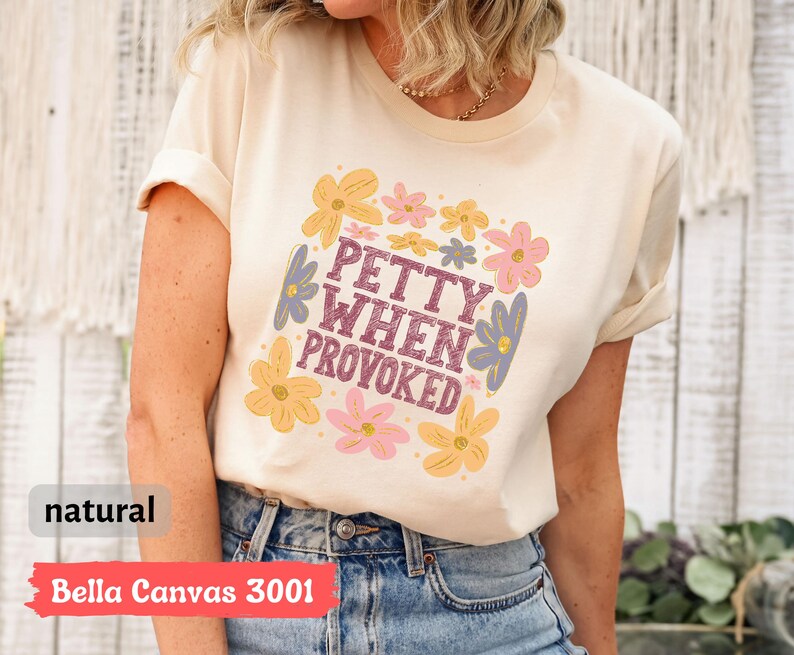 May include: A cream-colored t-shirt with the text "PETTY WHEN PROVOKED" in a square floral design. The design includes yellow, pink, and blue flowers. The shirt is made of Bella Canvas 3001 material.