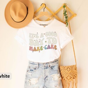 Peut inclure: T-shirt blanc avec le texte "It's a good day to make cake" en couleurs arc-en-ciel avec des &eacute;toiles. Le t-shirt est sur un cintre et a un chapeau marron et un sac tiss&eacute; &agrave; c&ocirc;t&eacute;.