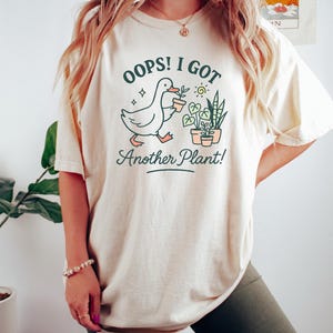 May include: Cream-colored t-shirt with the text "Oops! I Got Another Plant!" and a graphic of a duck carrying a potted plant. The design includes potted plants and a sun. The shirt is a casual, comfortable style.