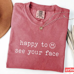 May include: A crimson red t-shirt with the text "happy to see your face" and a white smiley face graphic.