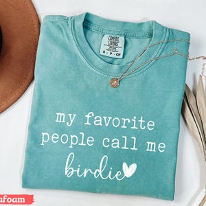 May include: Seafoam green t-shirt with the text "my favourite people call me birdie" in white. A gold necklace with a pendant rests on the shirt. A brown hat and dried plant are in the background.