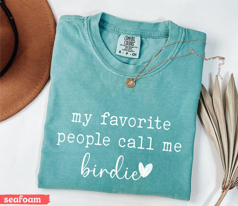 May include: Seafoam green t-shirt with the text "my favourite people call me birdie" in white. A gold necklace with a pendant rests on the shirt. A brown hat and dried plant are in the background.