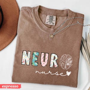 Puede incluir: Camiseta color espresso con las palabras "NEUR" en estampado floral y "nurse" en cursiva. Tambi&eacute;n presenta un gr&aacute;fico de cerebro con flores y un coraz&oacute;n. Un collar plateado con un colgante est&aacute; sobre la camiseta.