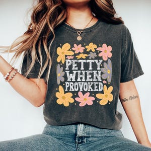 May include: Dark gray t-shirt with the text "PETTY WHEN PROVOKED" in a white, vintage-style font, surrounded by colorful flowers in shades of yellow, pink, and purple. The shirt is a casual, comfortable style.