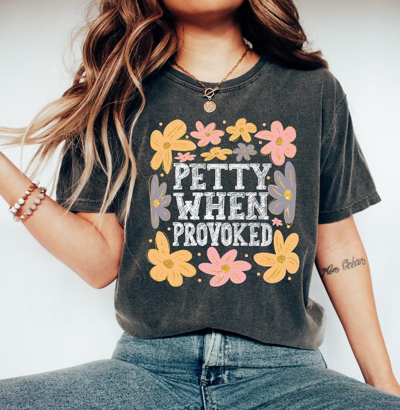 May include: Dark gray t-shirt with the text "PETTY WHEN PROVOKED" in a white, vintage-style font, surrounded by colorful flowers in shades of yellow, pink, and purple. The shirt is a casual, comfortable style.
