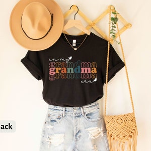 May include: Black t-shirt with the text "in my grandma era" in rainbow colors. The shirt is hanging on a wooden hanger with a straw hat and a woven bag.