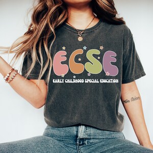 K&ouml;nnte beinhalten: Ein dunkelgraues T-Shirt mit einer bunten Grafik der Buchstaben "ECSE" im Retro-Stil. Die Buchstaben sind von kleinen wei&szlig;en Sternen umgeben. Unter der Grafik ist der Text "EARLY CHILDHOOD SPECIAL EDUCATION" in Wei&szlig; gedruckt.