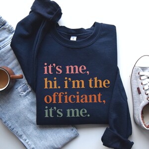 May include: A navy blue sweatshirt with the text "it's me, hi. i'm the officiant, it's me." printed in orange, pink, and green.