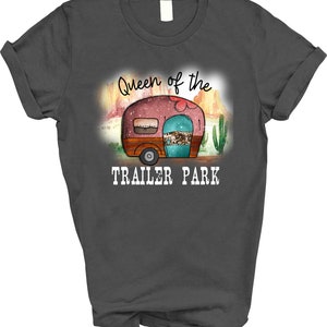 May include: A dark gray t-shirt with the text "Queen of the Trailer Park" in white. The design features a cartoon camper with a pink, turquoise, and brown color scheme.