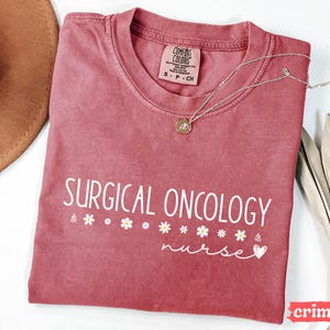 May include: A crimson red t-shirt with the text "SURGICAL ONCOLOGY nurse" in white lettering. The shirt has a distressed look and features a small white heart and a row of white flowers.