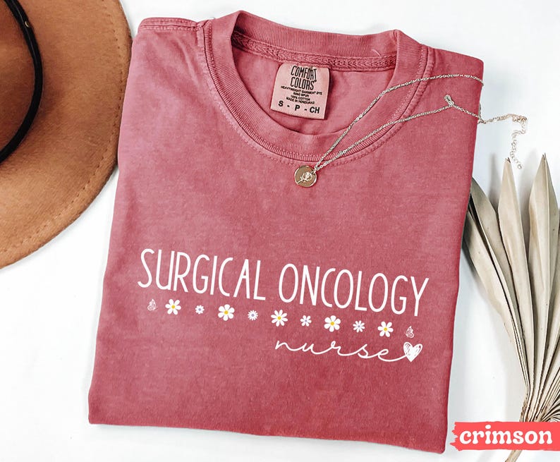 May include: A crimson red t-shirt with the text "SURGICAL ONCOLOGY nurse" in white lettering. The shirt has a distressed look and features a small white heart and a row of white flowers.